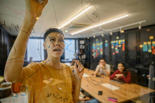 A woman writing on a clear board, brainstorming with a team.